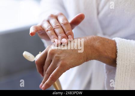 À la salle de bain à la maison, femme caucasienne mature portant une robe blanche, appliquant une lotion sur les mains Banque D'Images