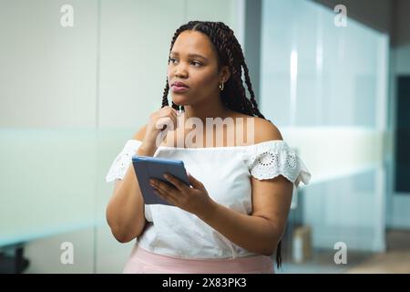 Au bureau moderne pour les affaires, jeune femme biraciale tenant tablette et pensant Banque D'Images