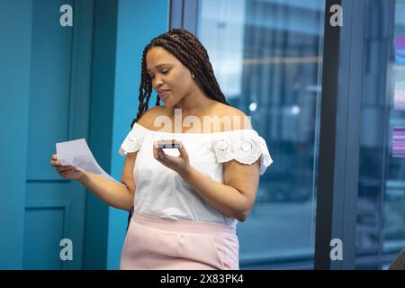 Jeune femme biraciale avec de longues tresses par bureau pour la fenêtre d'affaires, tenant du papier et une boîte Banque D'Images