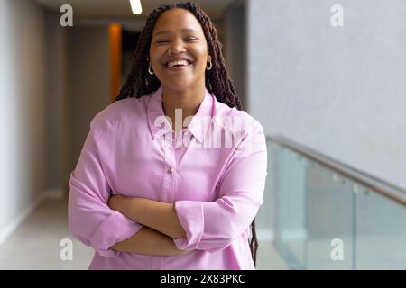 Au bureau moderne pour les affaires, jeune femme biraciale se tient souriante avec les bras croisés Banque D'Images