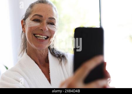 À la salle de bain à la maison, femme caucasienne mature avec crème pour le visage prenant selfie Banque D'Images