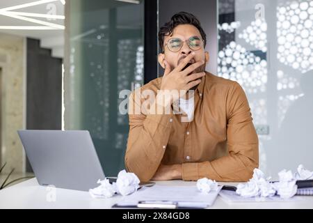 Homme d'affaires fatigué bâillant à son bureau entouré d'un ordinateur portable et de papiers froissés dans un cadre de bureau moderne. Concept de fatigue, surmenage et stress. Banque D'Images