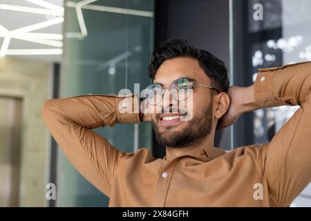 Jeune homme détendu avec des lunettes souriant en toute confiance dans un cadre de bureau moderne. Chemise marron, pose décontractée et fond contemporain. Banque D'Images