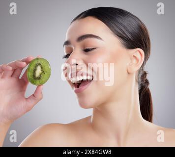 Beauté, visage et kiwi avec femme naturelle en studio sur fond gris pour l'alimentation, la santé ou la nutrition. Manger, nourriture ou fruits et modèle heureux avec frais Banque D'Images