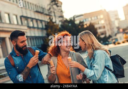 Groupe de touristes appréciant en vacances, jeunes amis s'amusant à marcher dans la rue de la ville manger de la crème glacée pendant le coucher du soleil. Banque D'Images