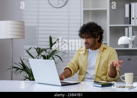 Jeune homme exprimant sa frustration tout en travaillant sur un ordinateur portable dans un environnement de bureau contemporain. La scène capture un moment stressant pendant une journée de travail. Banque D'Images