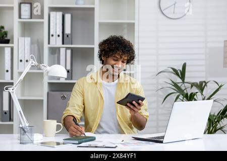 Jeune homme souriant travaillant à la maison avec une tablette et un ordinateur portable, tenant un stylo. Environnement de travail à distance détendu et productif avec fournitures de bureau et mobilier moderne. Banque D'Images