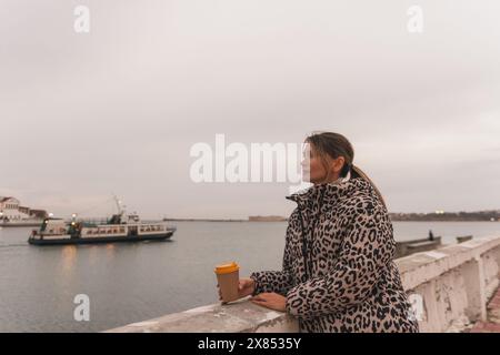 Une femme portant un manteau imprimé léopard se tient sur un rebord surplombant un plan d'eau. Elle tient une tasse de café et regarde l'eau. La scène Banque D'Images