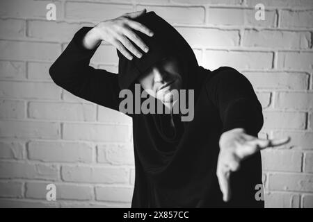 Un homme portant un sweat à capuche noir danse énergiquement avec des mouvements expressifs de la main devant un mur de briques blanches. Banque D'Images