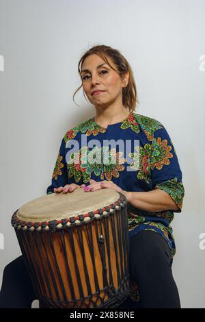 Une femme latine adulte portant un vêtement de style africain est assise jouant d'un tambour yembe regardant la caméra. Le tambour est en bois et a un et noir Banque D'Images