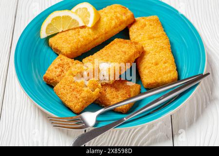 filet de pollock d'alaska pané croustillant avec couverts et tranches de citron sur plat bleu sur table en bois blanc, vue horizontale d'en haut Banque D'Images