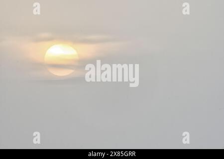 Une scène sereine du soleil partiellement obscurcie par des nuages doux au crépuscule, projetant une lueur chaude à travers le ciel brumeux. Banque D'Images