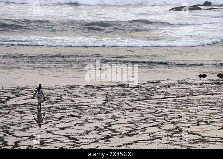 Une femme marche le long d'une plage avec des motifs de sable uniques créés par la marée qui recule. Les vagues douces en arrière-plan ajoutent à la sérénité et au mépris Banque D'Images