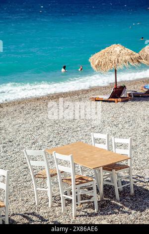 Table en bois avec quatre chaises en bois, sur la plage de galets de Loutraki, en Grèce, où les nageurs profitent des eaux Azur pendant les vacances d'été. Banque D'Images