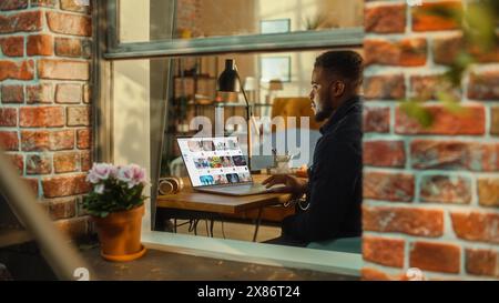 Homme noir utilisant un ordinateur portable avec affichage de plate-forme de streaming vidéo tout en étant assis derrière une table dans un salon élégant. Influenceur naviguant sur Internet. Prise de vue de l'extérieur de la fenêtre. Banque D'Images