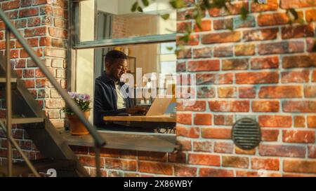 Jeune homme beau utilisant l'ordinateur portable, assis dans le salon dans l'appartement. Homme d'affaires afro-américain vérifiant les médias sociaux, souriant, répondant aux e-mails de la maison. Vue sur la fenêtre de l'appartement. Banque D'Images