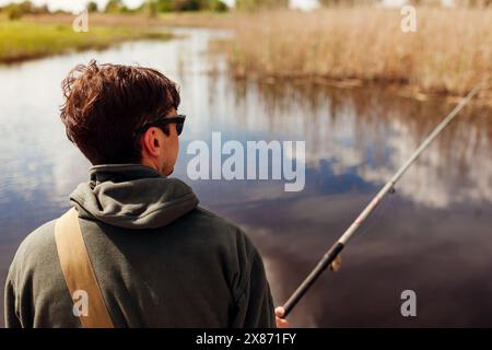 Vue arrière d'un jeune homme qui pêche portant un uniforme militaire. Gars assis sur le pont à travers la rivière tenant la tige. Concept de loisirs Banque D'Images