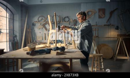 Portrait d'un charpentier mettant sur des lunettes, regardant un plan et commençant à assembler une chaise en bois. Concepteur de meubles professionnel travaillant dans un studio dans l'espace loft avec des outils sur les murs. Banque D'Images