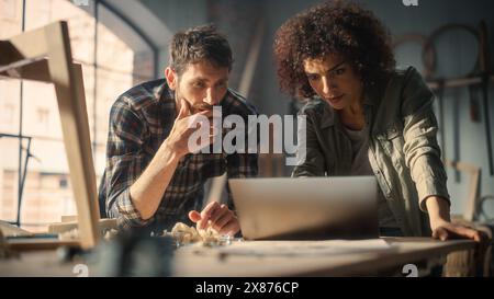 Excité jeune homme et femme utilisant un ordinateur portable, discutant d'un projet réussi dans un studio de menuiserie. Les concepteurs de meubles High Five et célèbrent la réception d'une occasion d'affaires de grande envergure. Banque D'Images