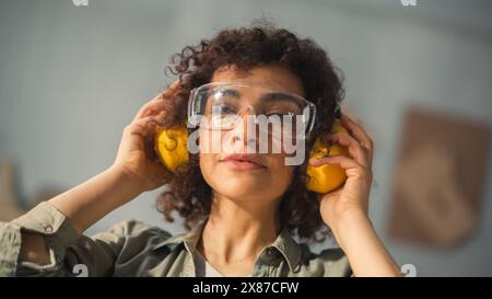 Portrait d'une belle femme multiethnique avec des cheveux bouclés et des yeux bruns mettant sur des lunettes de sécurité protectrices et casque antibruit de construction dans un environnement d'atelier. Banque D'Images