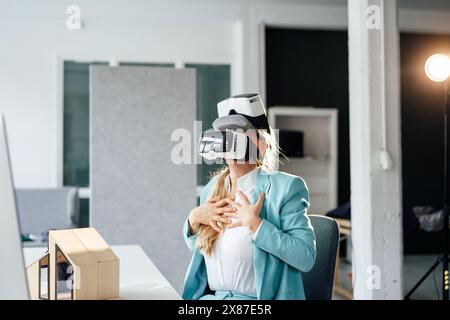 Femme d'affaires mature avec les mains sur la poitrine portant des casques de réalité virtuelle gestuelle au bureau Banque D'Images