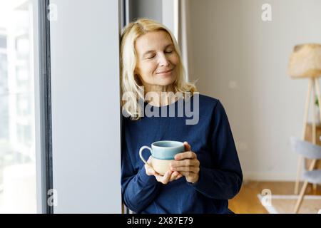 Femme souriante avec les yeux fermés tenant la tasse de thé par la fenêtre à la maison Banque D'Images