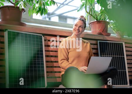 Femme d'affaires souriante utilisant un ordinateur portable assis jambes croisées au milieu des panneaux solaires dans le bureau Banque D'Images