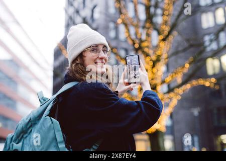Souriant beau touriste photographiant arbre décoré avec des lumières de chaîne dans la ville Banque D'Images