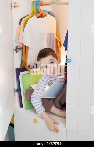 Garçon souriant assis les jambes croisées avec un livre dans la garde-robe à la maison Banque D'Images