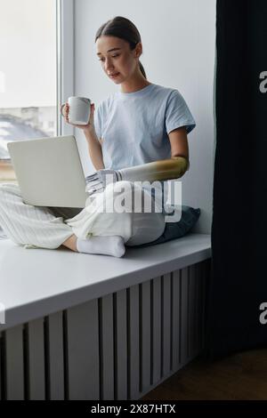 Jeune femme avec un bras prothétique buvant du café et utilisant un ordinateur portable sur le rebord de la fenêtre à la maison Banque D'Images