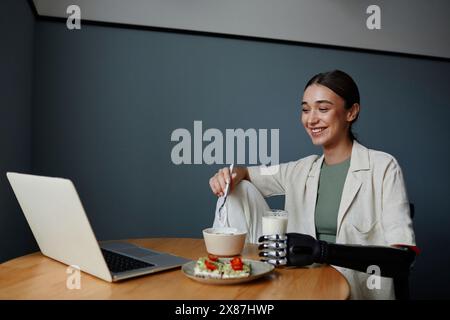 Femme heureuse avec le bras bionique noir manger un petit déjeuner sain et regarder la vidéo sur ordinateur portable à table Banque D'Images