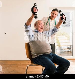 Médecin souriant debout à côté d'un homme âgé assis sur une chaise s'exerçant à la maison de retraite Banque D'Images