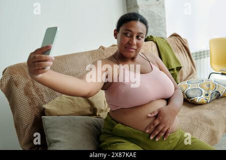 Femme enceinte de taille plus utilisant un téléphone intelligent pour prendre selfie assis sur le canapé à la maison Banque D'Images