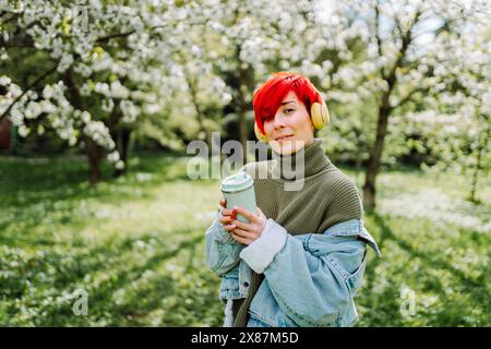 Femme rousse souriante écoutant de la musique avec une tasse réutilisable debout dans le parc Banque D'Images