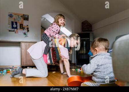 Fille souriante chevauchant sur le dos de la mère près du garçon jouant à la maison Banque D'Images