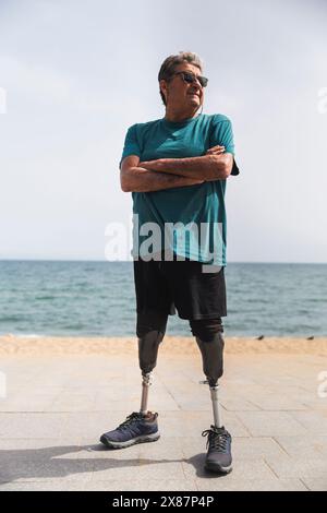 Homme âgé handicapé avec des jambes prothétiques debout avec les bras croisés à la plage le jour ensoleillé Banque D'Images