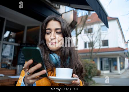 Femme tenant une cuillère dans la bouche et utilisant un téléphone intelligent près du café Banque D'Images