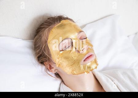 Belle fille blonde couchée dans le salon de spa avec les yeux fermés sous masque facial cosmétique doré Banque D'Images