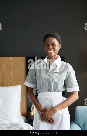 Portrait vertical d'une femme afro-américaine souriante en tant que femme de ménage portant un uniforme regardant la caméra dans la chambre Banque D'Images