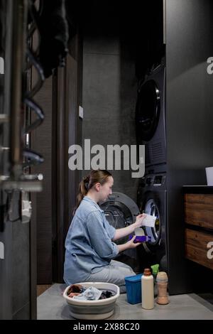 Femme assis devant une machine à laver chargeant des vêtements sales, serviettes pour laver des vêtements colorés. Banque D'Images