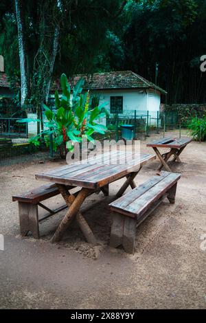 Plan vertical d'un ensemble de deux longues tables en bois avec des bancs de pique-nique extérieurs sur la surface non pavée dans un parc tropical avec une vieille maison modeste dans le Banque D'Images