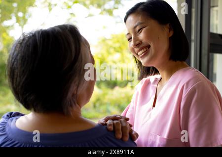 A la maison, jeune infirmière asiatique et patiente d'âge moyen, souriant en bleu et rose Banque D'Images