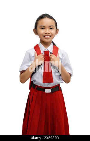 Portrait d'une jeune fille joyeuse en uniforme scolaire, se tenant avec confiance sur ses sangles de sac à dos, isolée sur un fond blanc avec espace de copie Banque D'Images