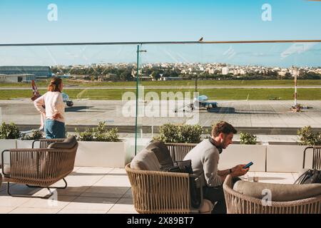 Voyageurs sur la terrasse extérieure du salon la Valette Club à l'aéroport de Malte. Un homme utilise son téléphone pendant qu'une femme surveille la piste Banque D'Images