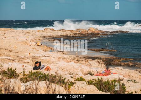 Bains de soleil se relaxant et lisant au soleil sur les rives rocheuses de Qawra point Beach, sont Paul's Bay, Malte Banque D'Images