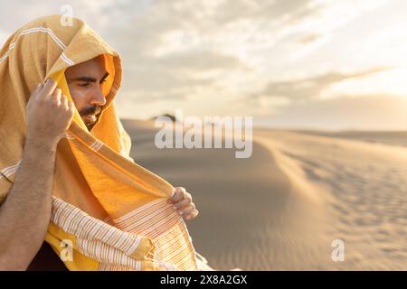 homme dans le désert en expédition avec son visage couvert contre la lumière au coucher du soleil regardant la caméra, dans la lumière dorée, parcourant le monde Banque D'Images