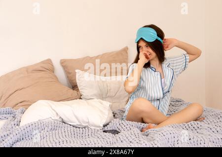 Adolescente vêtue d'un pyjama rayé se réveillant et enlevant le masque de sommeil dans le lit. Banque D'Images