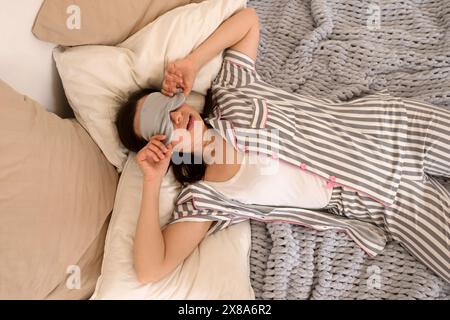 Femme en pyjama rayé gris et un masque de sommeil souriant ludique tout en tenant des oreillers sur une couverture en tricot. Banque D'Images