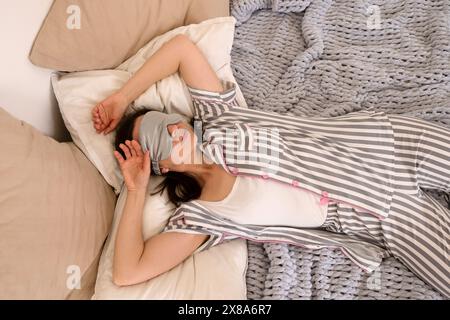 Femme en pyjama rayé gris et un masque de sommeil souriant ludique tout en tenant des oreillers sur une couverture en tricot. Banque D'Images