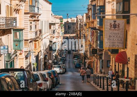 Une rue animée de la Valette présente une architecture charmante et bordée de voitures garées sous un ciel bleu clair Banque D'Images